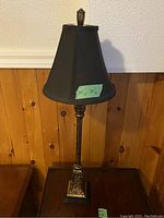 Tall floor lamp with ornate bronze-colored metal base and black tapered shade, placed against wood-paneled wall.