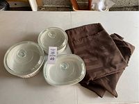 Three white CorningWare casserole dishes with glass lids stacked and set beside brown cloth napkins on white table