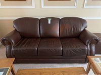 Front view of brown leather couch with three seat cushions and rolled arms showing nailhead trim detailing, placed in a room with beige walls and wooden furniture.