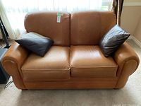Front view of tan leather loveseat with two pillows, on carpeted floor.