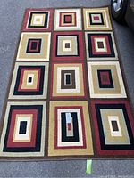 Full view of rectangular rug with repeating geometric squares and rectangles in multiple colors, placed on pavement with a car's tire partially visible.