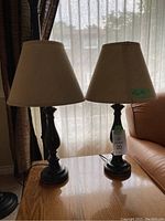 Photo of two matching table lamps side by side on wooden table, with beige fabric shades and turned dark bases.