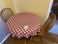 Round wooden table with checkered red and white tablecloth and two matching wooden chairs with vertical slat backrests.