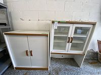 Two white cabinets side by side against a white wall, the one on the left with solid doors, the one on the right with glass doors and bottom shelf, placed on a gravel floor.