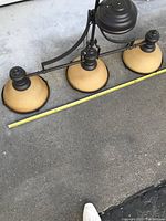 Three-light light fixture showing alabaster glass shades and iron frame with measuring tape stretched across length for size reference.
