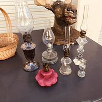 Five vintage oil lanterns of various glass types and styles on dark table with wicker basket and moose bust in background