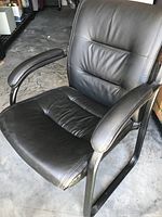 Black leather side chair with padded arms, metal sled base, and stitched detailing. Chair is positioned on concrete floor.