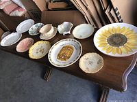 Overview of 10 vintage collector plates with varying shapes and designs on a wooden table including a large sunflower plate.