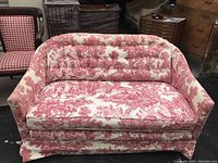 Front view of a vintage love seat upholstered in red and white toile fabric with scenic patterns, showing the overall style and condition.