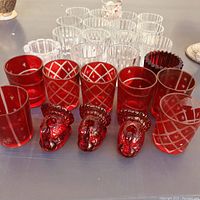 Photo showing 20 votive candle holders arranged in rows, featuring clear and red glass holders with patterns and three red rooster-shaped glass holders.