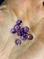 Hand holding a group of 10 small, round faceted amethyst gemstones with purple hues.