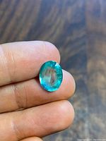 Front view of oval faceted blue-green gemstone held between fingers on wood background