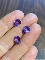 Three lab-created pear-shaped alexandrite gemstones held on fingertips showing color and size.