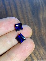Photo showing front faces of two square cut deep blue lab created sapphire gemstones held between fingers.