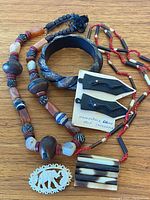 Vintage tribal jewelry collection on wooden surface showing multiple beaded necklaces, a carved bangle bracelet, a pendant, and ebony earrings on card.