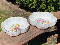 Two white lobed serving dishes with multi-colored floral patterns and gold trim seen outdoors on a wooden surface.