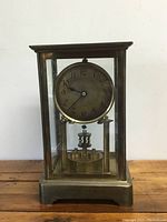 Full front view showing brass frame, glass panels, brass clock face with Arabic numerals, and pendulum mechanism inside.