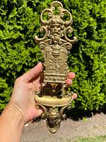 Front view of the antique ornate solid brass match holder held in hand showing detailed human face at the top and decorative scrolls.