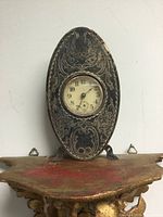 Front view of the antique small oval silver-plated clock showing etched floral patterns and the clock face with Arabic numerals and seconds dial.