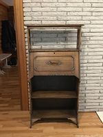 Front view showing the full secretaire cabinet against a light brick wall with the closed front desk panel and two open shelves below.