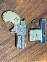 Photo of two vintage prop lighters on wooden surface showing Derringer style lighter with ornate silver-tone body and faux ivory grip, and rectangular lighter with mother-of-pearl panels.