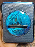 Front of metal case showing textured silver-tone surface with circular inlay of RMS Queen Mary ship on blue butterfly wing background.