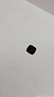 Small cushion cut red gemstone lying on white surface, showing size and color.