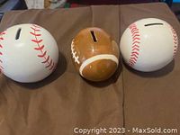 Three sports-themed ceramic piggy banks shaped as baseballs and a football, displayed on brown fabric.