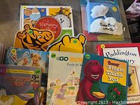 Photo of nine children's books showing covers including Nursery Rhymes with a tiger illustration, Baby Birds in the Sky stuffed animal packaging, Paddington's Nursery, and Barney Please Thank You book.