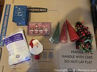 Photo showing various Christmas decorations including a blue metal candle holder with a reindeer design, a pack of fake snow, a Santa Claus fabric door hanger, a LOVE sign with four Christmas figurines, and four decorative Christmas trees in red and green.