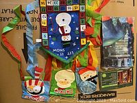Photo showing 3 colorful Christmas-themed windsocks laid out, including a penguin with Santa hat, Santa Claus face, and snowman, along with a snowman countdown calendar door hanger and packaging for the door cover with lights and sounds.