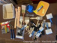 Set of painting tools and accessories spread on a brown surface including paint and art brushes, foam brushes, roller handles, paint cup, pour guard, tape, sponges, and stirrers.