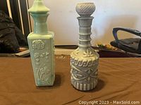 Photo of two vintage ceramic bottles on a brown surface, one square green with floral relief and one round gray with intricate raised designs and stopper.