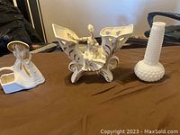 Three vintage decorative vases on a brown cloth surface: one white ceramic shaped as a woman with a hat, one hobnail white milk glass vase, and one white porcelain seated ballerina vase with cut-out floral designs.