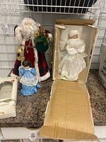 Photo of three Christmas tree toppers displayed on a surface against a wire grid backdrop: Santa Claus figure in plaid coat, small blue dress angel/doll, and white angel in box opened at front.
