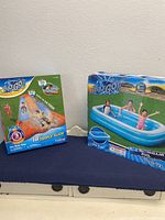 Both product boxes placed side by side on a blue surface against a wall. The left box is the 18ft triple slide water toy, the right box is the 8ft blue rectangular pool. Both boxes display images of children playing.