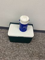 Green Coleman Model 5277 cooler with white lid, rectangular shape, placed on the floor.