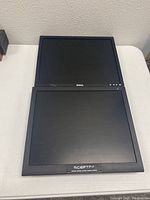 Photo showing two black flat screen monitors stacked, the top one Dell and the bottom one Sceptre, both powered off and on a white surface against a wall.