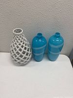 Three glass vases displayed upright on a flat surface against a white wall, showing white lattice vase and two blue striped vases