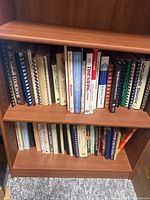 Two wooden shelves filled with over 50 cookbooks, mostly spiral-bound and some hardcover, arranged vertically