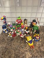 Photo showing a collection of M&M's character figurines and other assorted toy figures arranged on a countertop, with a mesh grid background.