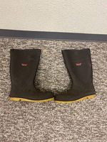 Side by side image of two black Tingley Profile water boots with yellow soles placed on a carpeted floor.
