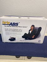 Box front showing Turbo Abs logo, tagline 'The Ultimate Abdominal Fitness System' and image of the workout chair with a model demonstrating use.