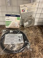 Lot contents showing Kenmore washing machine connector in unopened packaging and Pro-Dynamic dryer power cord in partially open packaging on countertop.