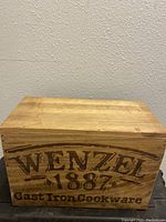 Front view of rectangular wooden box with 'Wenzel 1887 Cast Iron Cookware' branding burned on the front panel.