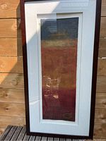 Photo showing the framed abstract art print with visible dark wood frame and white matting, displayed outdoors with natural light and some reflections.