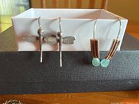 Pair of sterling silver dragonfly earrings and pair of earrings with blue round stones and brown elongated beads hanging on white box with a black surface background.