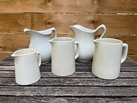 All five white jugs arranged on a wooden table showing size and shape variations