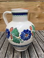 Front view of a ceramic jug showing hand-painted blue flowers, green leaves, and orange petals on white glaze, with handle and spout visible.