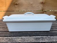 Side view of the white Le Creuset vegetable dish with lid on wooden surface showing rectangular shape and ridged side handles.